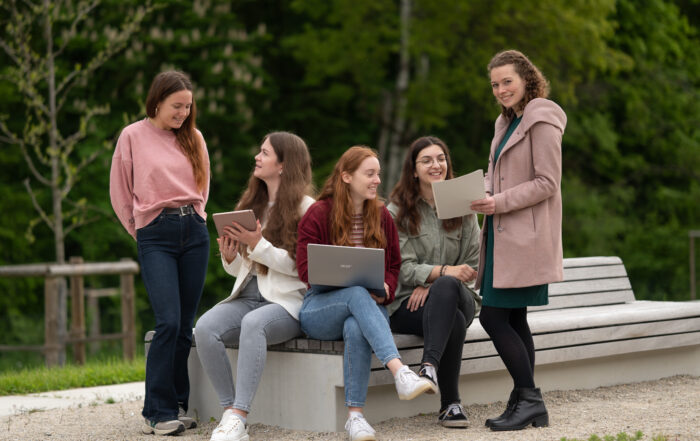 Fünf Frauen sitzen mit Laptops und Notizbüchern auf einer Bank im Freien an der Hochschule Coburg. Sie lächeln und unterhalten sich vor der üppig grünen Kulisse eines Parks.