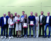Eine Gruppe von elf Personen steht vor einer hellgrünen Wand an der Hochschule Coburg. Drei Personen in der ersten Reihe zeigen stolz ihre Zertifikate, während eine andere Person einen Blumenstrauß hält. Alle sind in legerer Business-Kleidung gekleidet und vermitteln damit den Geist der Leistung und Einheit.