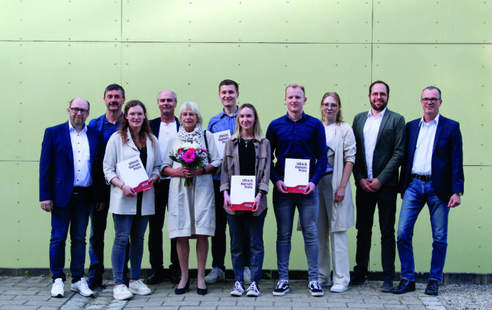 Eine Gruppe von elf Personen steht vor einer hellgrünen Wand an der Hochschule Coburg. Drei Personen in der ersten Reihe zeigen stolz ihre Zertifikate, während eine andere Person einen Blumenstrauß hält. Alle sind in legerer Business-Kleidung gekleidet und vermitteln damit den Geist der Leistung und Einheit.
