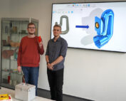 Zwei Personen stehen vor einem Bildschirm an der Hochschule Coburg, auf dem ein 3D-Modell einer Pfeife zu sehen ist. Ein Pfeil verbindet das Foto der echten Pfeife mit dem gerenderten Bild. Eine Person hält ein kleines rotes Objekt, während Regale mit verschiedenen Gegenständen einen faszinierenden Hintergrund bilden.