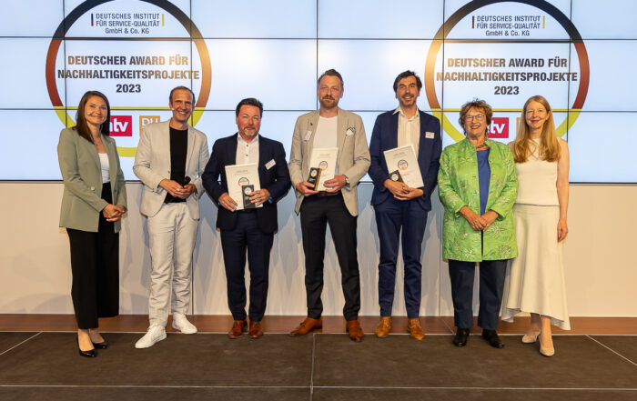 Eine Gruppe von Menschen, darunter Vertreter der Hochschule Coburg, steht lächelnd auf der Bühne, hält Auszeichnungen in den Händen. Hinter ihnen hängt ein mit Logos verziertes Banner mit der Aufschrift „Deutscher Award für Nachhaltigkeitsprojekte 2023“. Die Gruppe besteht aus drei Frauen und vier Männern in Business-Kleidung.
