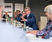Ein Mann im Anzug spricht und gestikuliert, während er an einem Konferenztisch sitzt, umgeben von Menschen, vermutlich an der Hochschule Coburg. Papiere, Flaschen und Mikrofone liegen verstreut auf dem Tisch herum, während die Teilnehmer in eine lebhafte Diskussion vertieft zu sein scheinen.