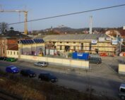 Straßenansicht einer Baustelle mit mehreren davor geparkten Fahrzeugen. In der Mitte steht ein großes Gebäude mit Gerüst, möglicherweise ein zukünftiger Flügel der Hochschule Coburg, mit einem Kran im Hintergrund. In der Ferne ist unter einem klaren Himmel eine Struktur auf einem Hügel zu erkennen.
