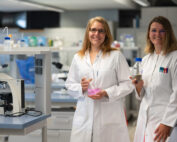 Zwei Wissenschaftler der Hochschule Coburg stehen lächelnd in Laborkitteln in einem modernen Labor. Einer hält einen Kolben mit rosa Flüssigkeit, der andere eine Flasche. Auf dem Tisch neben ihnen sind ein Mikroskop und elegante Laborgeräte zu sehen, die Innovation und Entdeckung symbolisieren.