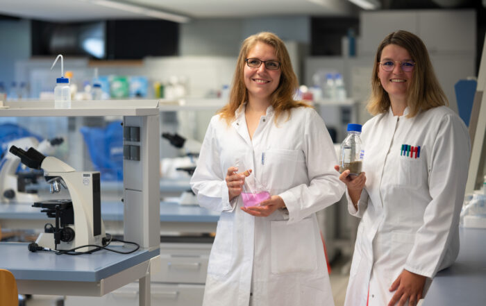 Zwei Wissenschaftler der Hochschule Coburg stehen lächelnd in Laborkitteln in einem modernen Labor. Einer hält einen Kolben mit rosa Flüssigkeit, der andere eine Flasche. Auf dem Tisch neben ihnen sind ein Mikroskop und elegante Laborgeräte zu sehen, die Innovation und Entdeckung symbolisieren.
