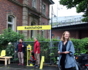 Eine lächelnde Frau steht neben einem gelben Schild mit einer Fahrradstation an der Hochschule Coburg. Hinter ihr stehen zwei Männer, einer benutzt die Pumpe und der andere hat ein Fahrrad. Im Hintergrund sind eine Holzbank und grünes Laub zu sehen, in der Nähe ein Backsteingebäude und eine Überführung.