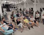 Eine Gruppe von Menschen der Hochschule Coburg sitzt in einem geräumigen, hellen Raum mit hoher Decke auf Liegestühlen und Sitzsäcken, lächelt und hebt die Hände. Einige benutzen Laptops, andere entspannen sich. Im Hintergrund sind Schreibtische und Bürogeräte zu sehen.