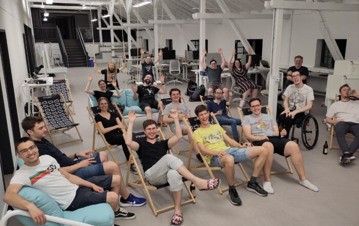 Eine Gruppe von Menschen der Hochschule Coburg sitzt in einem geräumigen, hellen Raum mit hoher Decke auf Liegestühlen und Sitzsäcken, lächelt und hebt die Hände. Einige benutzen Laptops, andere entspannen sich. Im Hintergrund sind Schreibtische und Bürogeräte zu sehen.