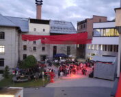 Diese Freiluftveranstaltung findet im Innenhof der Hochschule Coburg inmitten von Gebäuden im Industriestil statt. Über ihnen schwebt ein roter Baldachin, der einen warmen Schein auf die Menschenmenge wirft, die sich um Tische und eine Bühne mit leuchtend roter Beleuchtung versammelt hat. Der bedeckte Himmel verleiht der lebhaften Atmosphäre eine intime Note.