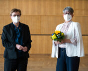 Zwei maskierte Personen stehen drinnen auf einem Holzboden in der Hochschule Coburg. Eine hält einen Strauß gelber und blauer Blumen in der Hand und trägt eine weiße Jacke und ein gepunktetes Hemd. Die andere trägt einen dunklen Anzug und eine blaugrüne Bluse. Beide haben kurze Haare und schauen in die Kamera.