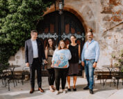 Eine Gruppe von fünf Personen steht lächelnd vor einem rustikalen Steingebäude mit Efeu, das an den charmanten Campus der Hochschule Coburg erinnert. Eine Person in der Mitte hält eine große runde Schachtel mit einem aufgedruckten Design. Zwei kleine runde Tische mit Stühlen stehen auf beiden Seiten der Gruppe.