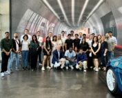 Eine bunt gemischte Gruppe von Menschen posiert gemeinsam vor einer abstrakt gestalteten Wand in der Hochschule Coburg und lächelt in die Kamera. Am rechten Rand ist ein blaues Auto angeschnitten zu sehen.