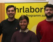 Drei Männer stehen vor einem gelb-schwarzen Schild der Hochschule Coburg. Sie lächeln und sind leger gekleidet, wobei der Hintergrund teilweise den Text des Schildes freigibt.