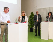 Vier Personen in formeller Kleidung nehmen an einer Konferenzdiskussion der Hochschule Coburg teil. Zwei stehen an Rednerpulten und sprechen, die anderen beiden hören aufmerksam zu. Auf den Rednern stehen Wasserflaschen, und eine Topfpflanze verleiht dem Hintergrund einen Hauch von Grün.
