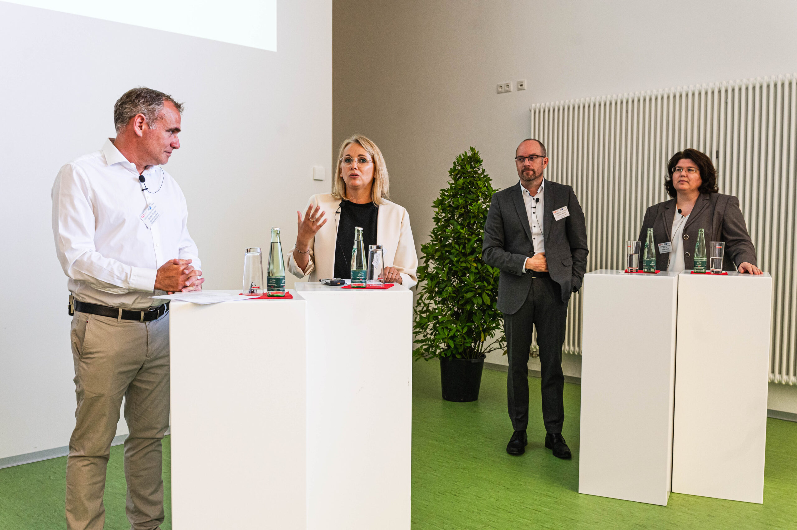 Vier Personen in formeller Kleidung nehmen an einer Konferenzdiskussion der Hochschule Coburg teil. Zwei stehen an Rednerpulten und sprechen, die anderen beiden hören aufmerksam zu. Auf den Rednern stehen Wasserflaschen, und eine Topfpflanze verleiht dem Hintergrund einen Hauch von Grün.