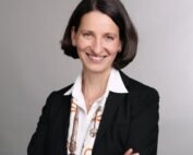 Eine Frau mit schulterlangem, dunklem Haar lächelt in die Kamera. Ihr schwarzer Blazer über einem weißen Hemd und ein Schal der Hochschule Coburg sind dazu getragen. Ihre Arme hat sie selbstbewusst vor dem grauen Hintergrund verschränkt.