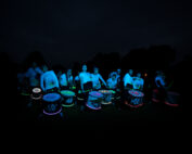 Eine Gruppe von Trommlern tritt nachts auf, beleuchtet durch leuchtende Neonlichter an ihren Trommeln und ihrer Kleidung. Ihre leuchtenden Farben heben sich vom dunklen Hintergrund ab und erzeugen eine lebendige und energiegeladene Szene, die an ein Festival der Hochschule Coburg erinnert.