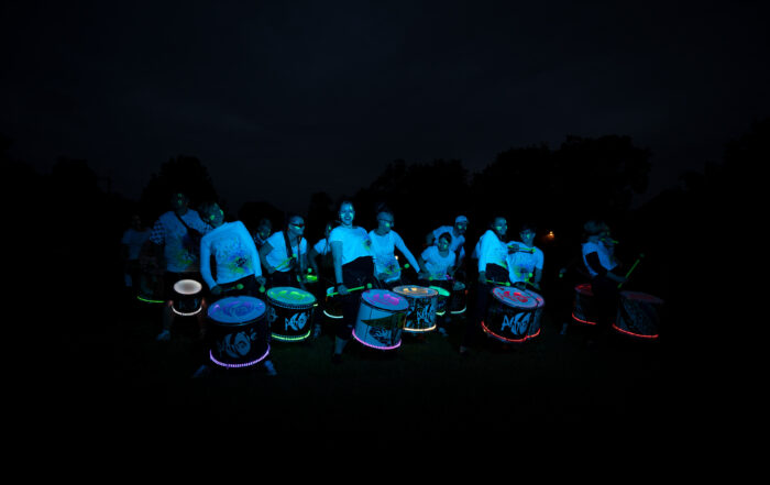 Eine Gruppe von Trommlern tritt nachts auf, beleuchtet durch leuchtende Neonlichter an ihren Trommeln und ihrer Kleidung. Ihre leuchtenden Farben heben sich vom dunklen Hintergrund ab und erzeugen eine lebendige und energiegeladene Szene, die an ein Festival der Hochschule Coburg erinnert.