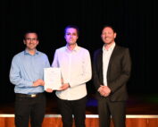 Drei Männer stehen auf einer Bühne. Der Mann links im blauen Hemd hält stolz ein Zertifikat der Hochschule Coburg in den Händen, der Mann in der Mitte trägt ein weißes Hemd. Der Mann rechts trägt einen schwarzen Anzug und lächelt in die Kamera. Im Hintergrund ist ein dunkler Vorhang zu sehen.