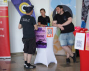 Drei Menschen unterhalten sich an einem Tisch mit Flyern und einem QR-Code. Zwei Männer in schwarzen Hemden unterhalten sich mit einer Frau dazwischen. Im Hintergrund sind die rot-bunten Banner der Hochschule Coburg mit unterschiedlichen Texten und Logos zu sehen.