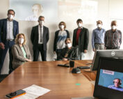 Eine Gruppe von acht Personen mit Masken steht und sitzt um einen Konferenztisch in dem hellen Raum der Hochschule Coburg. Auf zwei Computermonitoren werden Videoanrufe angezeigt, an der Wand dahinter wird eine Projektion abgespielt.