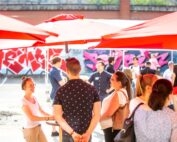 Eine Gruppe von Menschen, darunter Studierende der Hochschule Coburg, trifft sich unter roten Sonnenschirmen im Freien. Im Hintergrund ist eine mit Graffiti bedeckte Wand zu sehen. Einige Leute stehen und unterhalten sich, während andere sich um Tische herum versammeln. Die Atmosphäre ist locker und fröhlich.