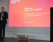 Ein Mann im Anzug präsentiert mit Mikrofon auf der Bühne der Hochschule Coburg. Hinter ihm läuft auf einer großen Leinwand ein Vortrag mit dem Titel „Pflege-Coop! Wege aus der Pflegefalle“ über ein innovatives Pflegekonzept von Prof. Dr. Eberhard Nofer vom 23.4.2024.