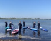 Eine Gruppe von sieben Personen in Neoprenanzügen steht auf Paddleboards auf einem ruhigen Gewässer unter einem klaren blauen Himmel. Im Hintergrund rahmen ein grasbewachsenes Ufer und eine Windturbine diesen malerischen Ort in der Nähe der Hochschule Coburg sanft ein.