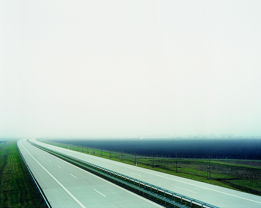 Unweit der Hochschule Coburg erstreckt sich eine lange, leere Autobahn unter nebligem Himmel. Grünes Gras säumt die Straße, flankiert von Feldern. Die Atmosphäre ist ruhig und trostlos, der Nebel verdeckt den Horizont.