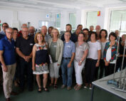 Eine Gruppe Erwachsener steht in einem hell erleuchteten Raum der Hochschule Coburg zusammen und lächelt in die Kamera. Sie befinden sich in einer Unterrichts- oder Werkstattumgebung, im Hintergrund sind Tische und verschiedene Geräte zu sehen.