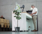 Eine Person steht an einem Rednerpult im Hörsaal der Hochschule Coburg und hält einen Vortrag. Auf dem Boden daneben steht eine kleine Topfpflanze, die in durchsichtige Plastikfolie eingewickelt ist. Im Hintergrund ist das Publikum teilweise zu sehen, das in Reihen von holzgetäfelten Sitzen sitzt.