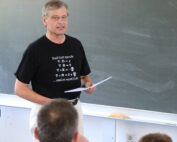 Ein Redner steht vor einer Tafel, hält Notizen in der Hand und trägt ein schwarzes T-Shirt mit Text und Symbolen. Im Vordergrund hören mehrere Personen aufmerksam zu. Die Kulisse lässt auf ein akademisches Umfeld schließen, möglicherweise in Verbindung mit der Hochschule Coburg.