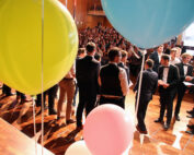Eine große Gruppe formell gekleideter Menschen versammelt sich in einem Saal mit Holzboden in der Hochschule Coburg. Bunte Luftballons in Gelb, Rosa und Blau tragen zur festlichen Atmosphäre bei und deuten auf ein besonderes Ereignis oder eine Zeremonie hin, an der viele teilnehmen.