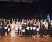 Auf einer Holzbühne steht eine große Gruppe von Menschen in Abendgarderobe und hält stolz Zertifikate oder Auszeichnungen der Hochschule Coburg in den Händen. Vor einem dunklen Vorhang im Hintergrund sind sie in Reihen aufgestellt.