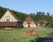 Eine ländliche Szene zeigt traditionelle Fachwerkhäuser mit steilen Dächern, umgeben von üppigem Gras und dichten Bäumen. Im Vordergrund liegt ein Stapel geschnittener Baumstämme unter einem klaren blauen Himmel, während sich eine schmale Straße in Richtung des Dorfes in der Nähe der ruhigen Landschaft der Hochschule Coburg schlängelt.