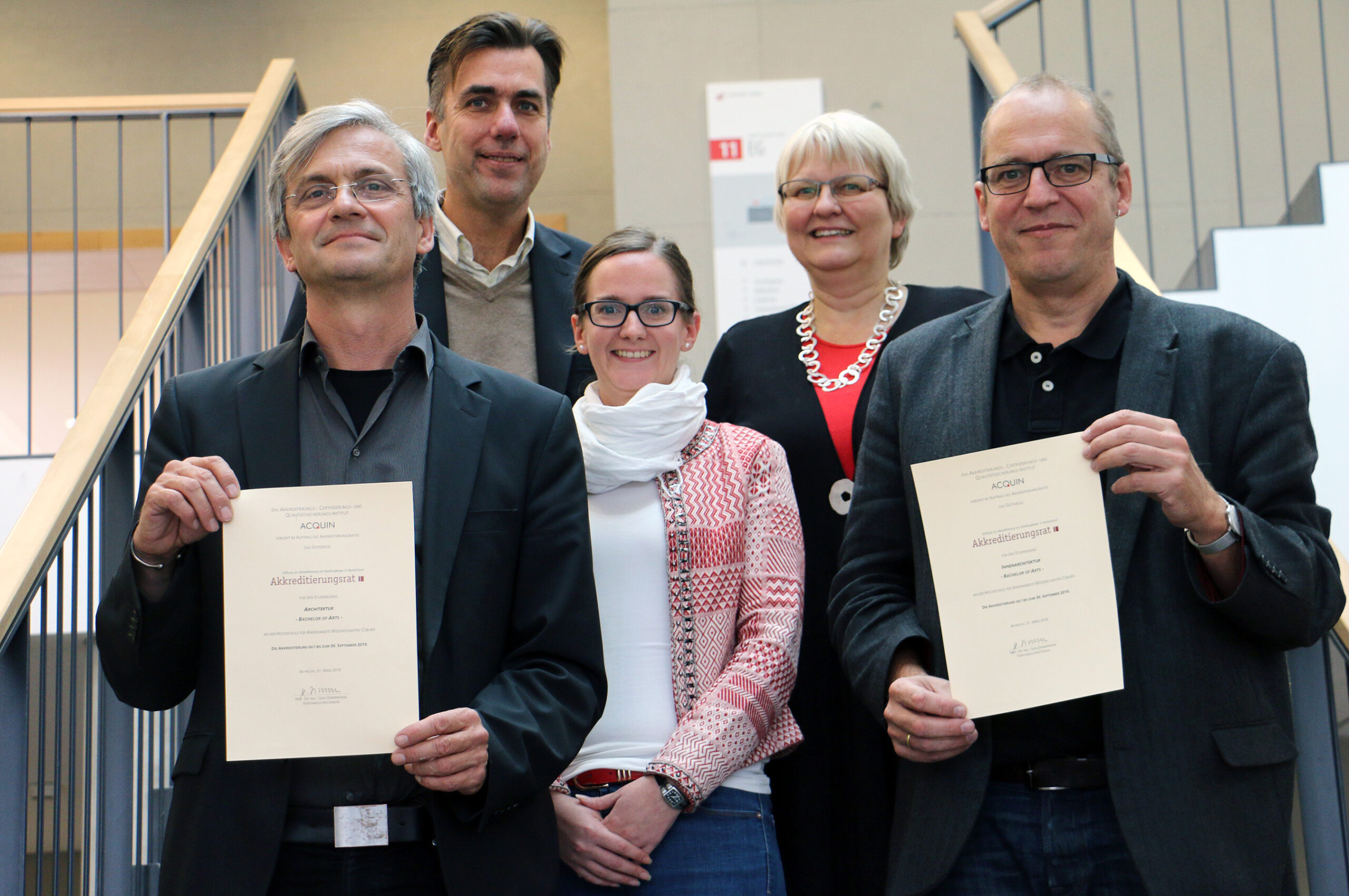 Fünf Personen stehen lächelnd zusammen und halten Zertifikate der Hochschule Coburg in den Händen. Zwei Personen in der ersten Reihe zeigen ihre Zertifikate prominent. Sie befinden sich in einem modernen Interieur mit einer Treppe und einem Geländer im Hintergrund.