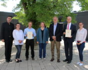 Eine Gruppe von sieben Personen steht im Freien vor einem Baum und einem Gebäude der Hochschule Coburg. Drei Personen halten stolz ihre Zertifikate in der Hand und sind in Business-Casual- bzw. Abendkleidung gekleidet.