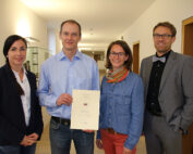 Vier Personen stehen zusammen in einem Flur der Hochschule Coburg. Der Mann in der Mitte hält ein Zertifikat in der Hand. Sie sind in Business-Casual-Kleidung gekleidet und lächeln in die Kamera.
