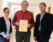 Drei Menschen stehen lächelnd in einem Flur der Hochschule Coburg. Die Person in der Mitte hält stolz ein Zertifikat in den Händen. Die Gruppe scheint eine akademische Leistung oder Auszeichnung zu feiern.