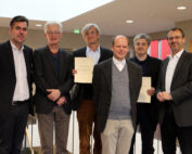 Sechs Männer in Anzügen stehen drinnen zusammen, zwei halten stolz ihre Zertifikate in den Händen. Sie stehen auf einem Treppenabsatz vor einem hellen Hintergrund in den Innenräumen der Hochschule Coburg. Alle lächeln, was auf einen feierlichen oder formellen Anlass schließen lässt.