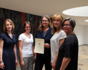 Fünf Frauen stehen in einem Raum der Hochschule Coburg zusammen. Eine hält ein Zertifikat in der Hand. Sie lächeln alle, im Hintergrund eine gemusterte Wand und darüber runde Deckenleuchten.