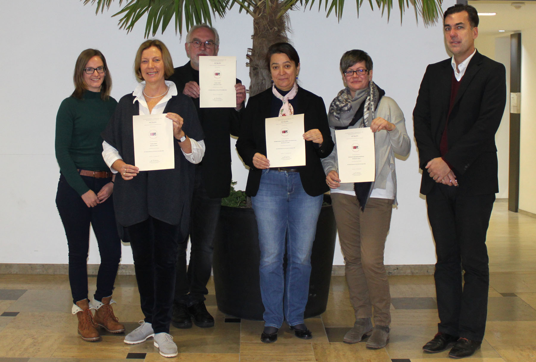 Sechs Personen stehen drinnen, fünf davon stolz mit Zertifikaten und einem Lächeln als Repräsentanten der Hochschule Coburg. Sie stehen vor einer großen Topfpflanze vor einer weißen Wand und schaffen so eine entspannte und lockere Atmosphäre.