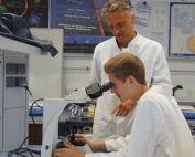 In einem mit wissenschaftlichen Postern geschmückten Labor an der Hochschule Coburg blickt ein junger Mann im Laborkittel durch ein Mikroskop, während ein älterer Mentor neben ihm steht und ihm Anleitung gibt.