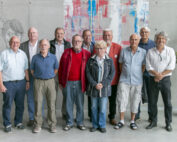 Eine Gruppe von elf älteren Erwachsenen, allesamt Männer, steht lächelnd in einer Reihe an der Hochschule Coburg. Sie befinden sich in einem modernen Ambiente mit grauen Wänden, an denen künstlerische Zeichnungen und ein farbenfrohes abstraktes Gemälde hängen. Auf der rechten Seite steht eine Topfpflanze.