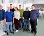 Eine Gruppe von sieben Personen, vier Männer und drei Frauen, steht lächelnd in einem gut beleuchteten Labor der Hochschule Coburg, im Hintergrund stehen Schreibtische und Geräte. In legerer Kleidung strahlen sie in dieser lebhaften akademischen Umgebung Professionalität und Freundlichkeit aus.
