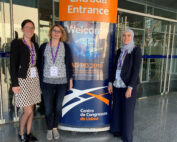 Drei Frauen stehen vor einem Konferenzplakat im Lissabonner Kongresszentrum und tragen stolz ihre Hochschulausweise der Hochschule Coburg. Sie lächeln herzlich in die Kamera, während das Plakat hinter ihnen die Teilnehmer der „AD/PD 2019“ in mehreren Sprachen begrüßt.