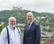 Zwei ältere Männer stehen draußen vor einem malerischen Hügel und einer Burg im Hintergrund und verkörpern den Charme der Hochschule Coburg. Einer trägt ein hellblaues Hemd mit Hosenträgern, der andere einen Anzug mit gestreifter Krawatte. Beide lächeln herzlich in die Kamera.