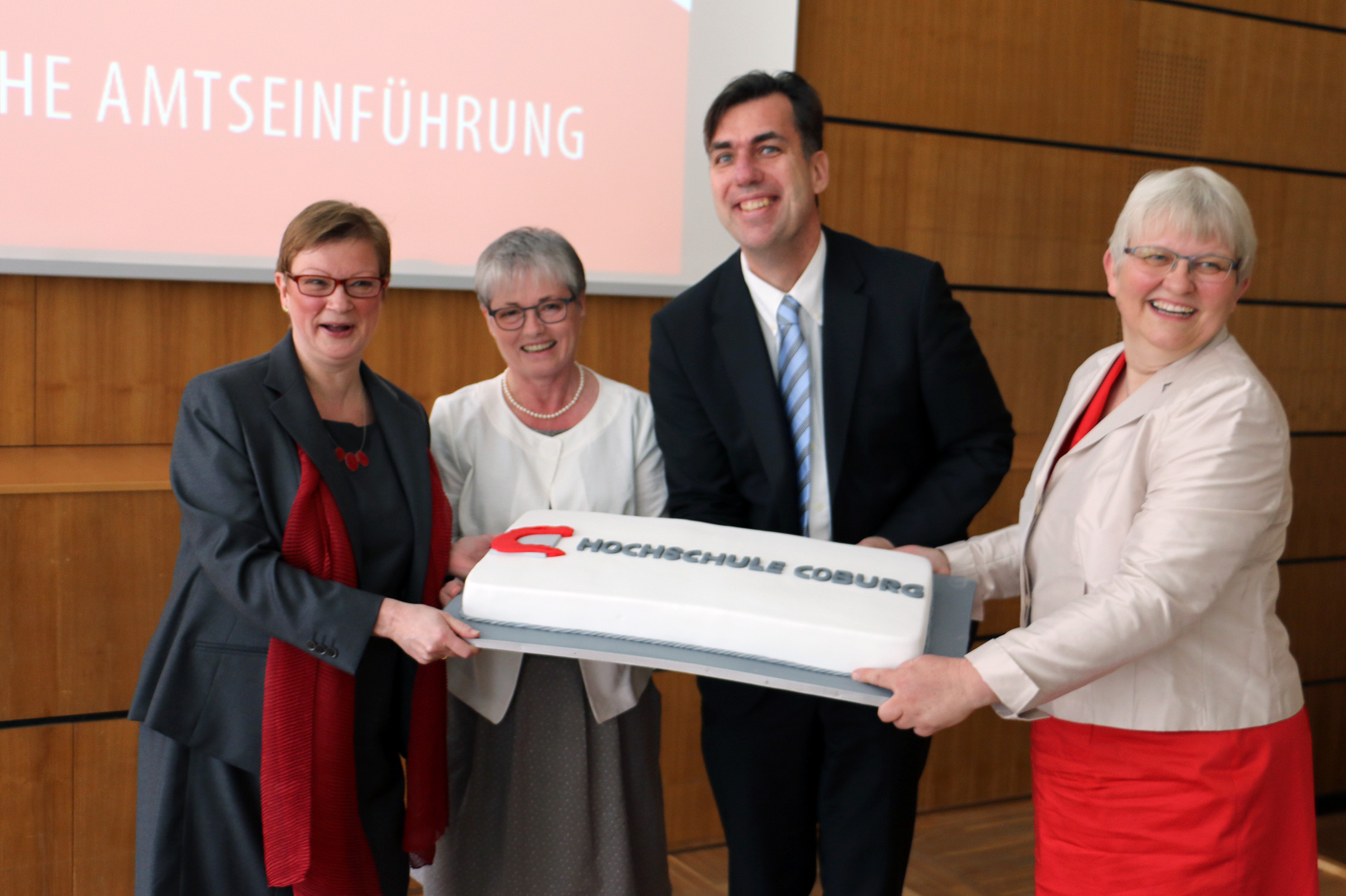 Vier Personen von der Hochschule Coburg lächeln und halten eine große rechteckige Torte mit dem Logo der Hochschule in den Händen. Stolz stehen sie vor einer Holzwand, über der teilweise eine Präsentationsfolie zu sehen ist.