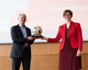Zwei Menschen stehen nebeneinander und halten jeweils einen Pokal in der Hand, der einen Fußball auf einem Holzsockel zeigt. Der Mann links trägt einen Anzug, die Frau rechts ein rotes Outfit. Hinter ihnen zeigt eine Projektionsleinwand einen Text auf Deutsch, der auf eine Veranstaltung an der Hochschule Coburg aufmerksam macht.