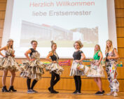 Auf der Bühne stehen sechs Personen in Kleidern aus Zeitungspapier. Hinter ihnen wird auf einem Bildschirm eine Begrüßungsnachricht auf Deutsch für die neuen Studierenden der Hochschule Coburg angezeigt. Die Kulisse scheint ein Auditorium oder ein Hörsaal zu sein, in dem die Menschen vor Erwartung und Aufregung über das bevorstehende akademische Leben summen.